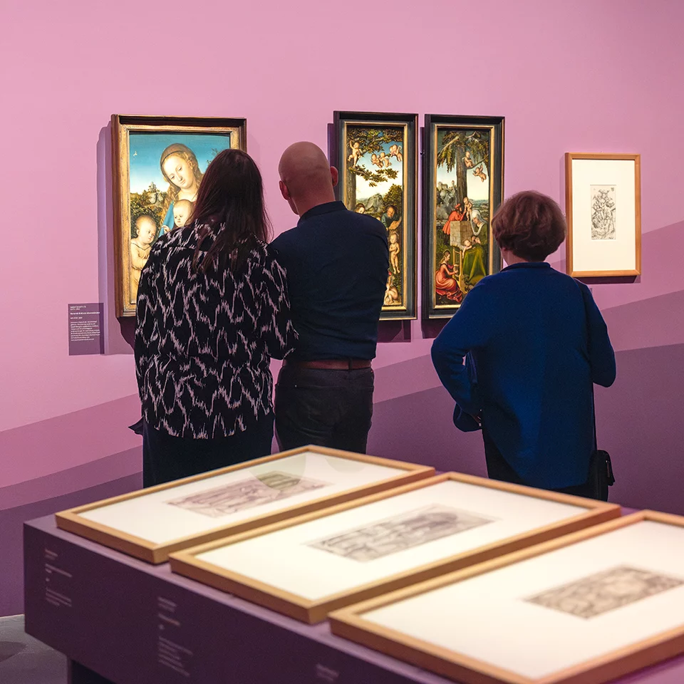 Visitors admire framed artworks in a gallery with a vibrant purple wall and displayed sketches in the foreground.